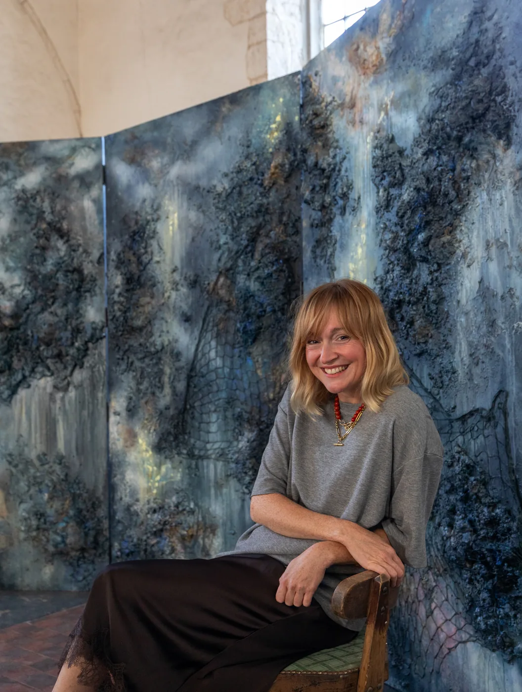 Tall storm-painted panels standing against the plastered wall of a medieval church interior, a woman seated in front of them