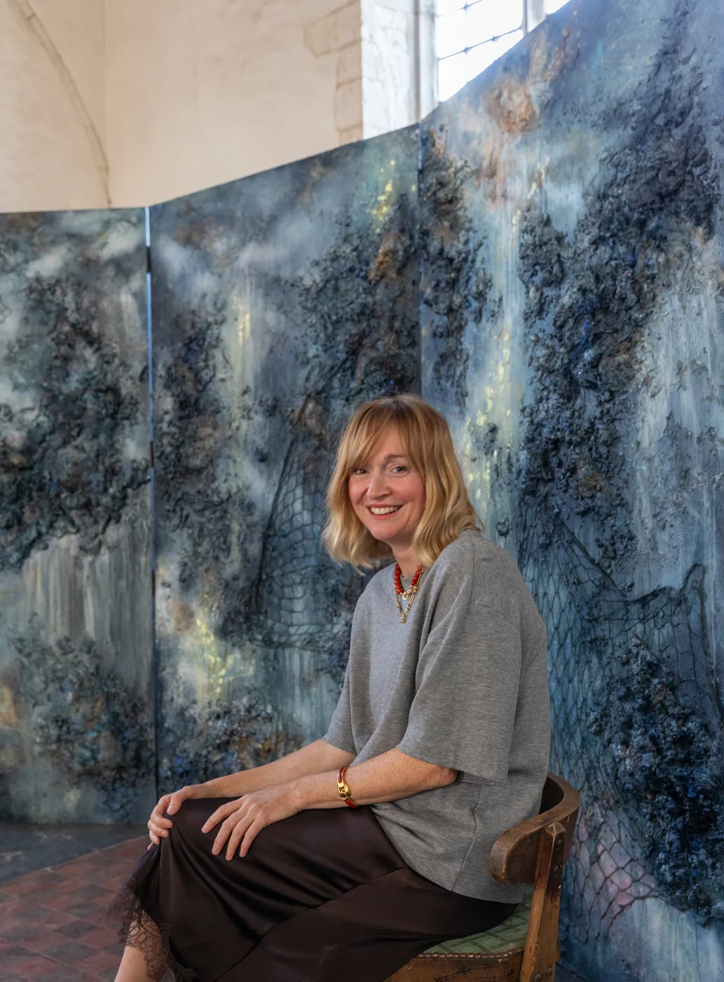 A woman sits on a wooden chair in front of tall painted panels in blue, grey and gold inside a white-walled church